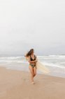 Весела молода спортсменка в купальнику з дошкою для серфінгу дивиться на піщане узбережжя проти штормового океану — стокове фото