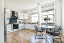Geräumige helle Küche mit weißen Schränken und Essecke mit weißem Tisch und Stühlen am Fenster — Stockfoto