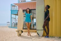 Contenuto giovani sportivi multirazziali con longboard e tavola da surf in piedi sulla riva sabbiosa contro la costruzione sotto il cielo nuvoloso — Foto stock