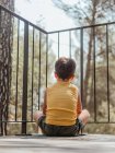 Rückansicht eines kleinen Jungen, der im Sommer auf der Veranda eines modernen Ferienhauses im Wald sitzt — Stockfoto