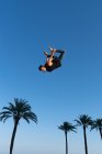 Vista lateral de ángulo bajo del atleta masculino activo haciendo backbend mientras que salta contra palmeras bajo cielo azul en la luz del sol - foto de stock