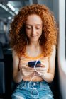 Interested woman with curly hair in ripped jeans text messaging on cellphone during trip on train in daytime — Stock Photo