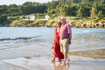 Sorrindo descalço casal de idosos em óculos de sol em pé na praia de areia molhada e desfrutando de dia ensolarado — Fotografia de Stock
