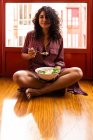 Молодая женщина с вьющимися волосами сидит, глядя на камеру со скрещенными ногами и ест супер миску с куриным горошком нарезанные яйца брокколи — стоковое фото
