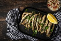Top view of tasty fried anchovies with juicy lemon piece and chopped parsley on tray against savory sauce — Stock Photo