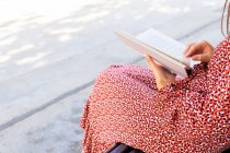 Обрезанная неузнаваемая женщина в стильной одежде сидит с открытой книгой на деревянной скамейке напротив здания со светлой стеной в дневное время — стоковое фото