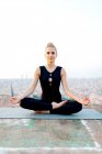 Full body of slim barefoot female in sportswear sitting in Ardha Padmasana with zen gesture during meditation on rooftop in evening — Stock Photo