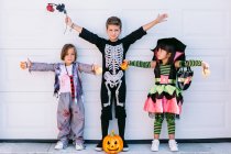 Fröhliche kleine Freunde in verschiedenen Halloween-Kostümen mit Kürbis und Accessoires heben die Arme und schauen in die Kamera, während sie an der weißen Wand zusammen stehen — Stockfoto