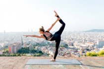 Seitenansicht einer flexiblen jungen Frau in Sportbekleidung, die in Dandayamana Dhanurasana-Pose während einer Yoga-Übung auf Matte in der Stadt steht — Stockfoto