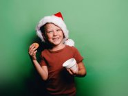 Menino sorridente adorável com chapéu de Papai Noel de Natal tomando biscoito de xícara contra fundo verde olhando para a câmera — Fotografia de Stock