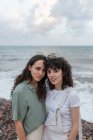 Jóvenes lesbianas novias en ropa casual abrazando mientras mira la cámara en la costa del océano bajo el cielo nublado - foto de stock