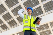 Baixo ângulo do engenheiro masculino hispânico de meia idade em uniforme com tablet de pé olhando para longe sob a central de energia solar — Fotografia de Stock