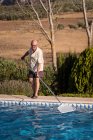 Corpo inteiro de homem idoso em óculos limpeza de água na piscina com esfregão no quintal — Fotografia de Stock