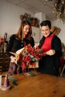 Alegres amigas de pie a la mesa con velas y haciendo creativos ramos de Navidad para la celebración de las fiestas - foto de stock