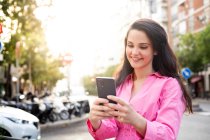Smiling fêmea em vestido de pé na calçada e mensagens de texto no celular — Fotografia de Stock