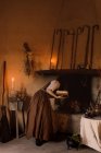 Strega in abito lungo accendere un fuoco tenendo il suo libro magico di incantesimi in mano mentre in piedi in camera accogliente con scopa e calderone — Foto stock
