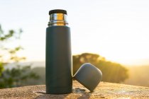 Modern stainless steel and plastic thermos with cup on rough surface under shiny sky at sundown — Stock Photo