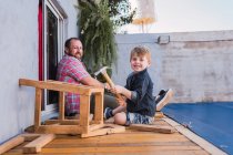 Barbudo pai ensinando filho com martelo trabalhando com madeira enquanto sentado no calçadão no fim de semana — Fotografia de Stock