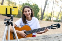 Fröhliche Teenie-Bloggerin spielt Akustikgitarre, während sie im Park ein Handy-Video auf Stativ aufnimmt — Stockfoto
