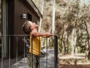 Вид сбоку маленького мальчика, стоящего на веранде современной кассы, расположенной в лесу летом — стоковое фото