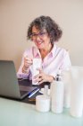 Содержание пожилая женщина предприниматель в наушниках, показывающих увлажняющий крем бутылку на камеру во время видеозвонка на ноутбуке в офисе — стоковое фото