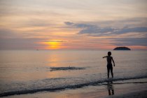 Vista posteriore piena lunghezza del ragazzo irriconoscibile in piedi sulla spiaggia di sabbia bagnata lavata dal mare al tramonto in Malesia — Foto stock