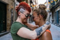 Vista lateral de pareja lesbiana joven de moda con tatuajes en gafas de sol abrazándose mirándose en el momento del beso en la ciudad - foto de stock