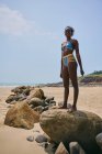 Vue latérale du contenu jeune femme afro-américaine en maillot de bain debout sur un rocher tout en regardant loin — Photo de stock