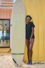 Fröhliche junge afroamerikanische Sportlerin in Bikini und T-Shirt blickt in einer Strandbar an der Küste mit Surfbrett in die Kamera — Stockfoto