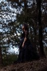 Знизу містична відьма в довгій чорній сукні і з пофарбованим обличчям, що стоїть, дивлячись в темні похмурі ліси — стокове фото
