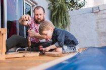 Bearded dad teaching son with hammer working with wood while sitting on boardwalk on weekend — Stock Photo