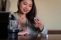 Урожай усміхнений молода етнічна жінка кладе кавовий стручок у виробника на стіл на домашній кухні — стокове фото