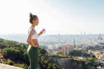 Side view of thoughtful slim female athlete with hand on hip and closed eyes drinking water from bottle during break from workout in town — Stock Photo