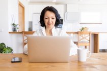 Joyeuse femme mature pigiste naviguant sur Internet sur netbook travaillant sur un nouveau projet tout en étant assise à table à la maison — Photo de stock