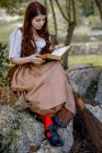 Hembra enfocada leyendo libro de magia mientras está sentada en la roca cerca de la escoba en el bosque - foto de stock