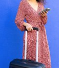 Cortado fêmea irreconhecível em vestido vermelho longo de pé com bagagem, enquanto a mensagem tet no smartphone na rua contra a parede azul durante o dia — Fotografia de Stock