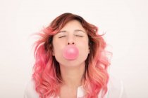 Веселая женщина с розовыми волосами выдувая жвачку пузыря и глядя прямо вперед с закрытыми глазами на светлом фоне — стоковое фото