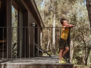 Vista lateral do menino em pé na varanda da casa de campo moderna localizada na floresta no verão — Fotografia de Stock