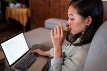 Seitenansicht einer interessierten jungen ethnischen Mitarbeiterin, die zu Hause auf dem Sofa am Netbook gegen Kaffee und Smartphone arbeitet — Stockfoto