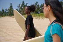 Lächelnde schwarze Sportlerin mit Longboard gegen asiatische Freundin mit Surfbrett, die sich im Meer unter wolkenlosem blauen Himmel freut — Stockfoto