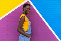 Vista laterale di felice giovane afroamericana sorridente mentre in piedi su parete luminosa colorata — Foto stock