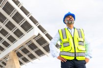 De baixo de empreiteiro masculino étnico maduro em hardhat e colete respirando com os olhos fechados enquanto está perto da central de energia solar — Fotografia de Stock