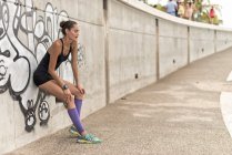 Vista lateral do corredor feminino atlético positivo em sportswear inclinado na parede ao fazer uma pausa durante o treino na cidade — Fotografia de Stock