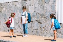 Schoolboy with backpack speaking with female friends while standing on tiled pavement against stone wall in sunlight — Stock Photo