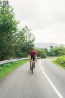 Deportista en bicicleta de montar casco de protección durante el entrenamiento en la carretera de asfalto contra la colina verde y los árboles bajo el cielo claro - foto de stock