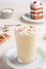 Glas Milchpunsch mit Zimtpulver auf geschlagenem Eiweiß gegen Kuchenstück auf Cafeteria-Tisch auf hellem Hintergrund — Stockfoto