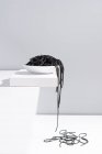 Estudio minimalista con espaguetis de tinta de calamar negro cayendo desde un tazón de cerámica completo sobre una mesa blanca - foto de stock