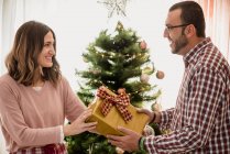 Cheerful adult female passing present box to astonished male beloved while celebrating New Year holiday in house — Stock Photo