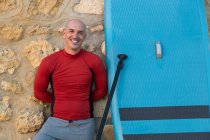 Surfista macho feliz em roupa de mergulho e chapéu de pé olhando para a câmera com remo e placa SUP enquanto se prepara para surfar na costa inclinada na parede de pedra — Fotografia de Stock