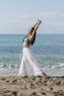 Vista lateral de la mujer étnica inclinada hacia atrás mientras está de pie en la postura de Ashta Chandrasana durante la práctica de yoga en la costa arenosa contra el océano - foto de stock